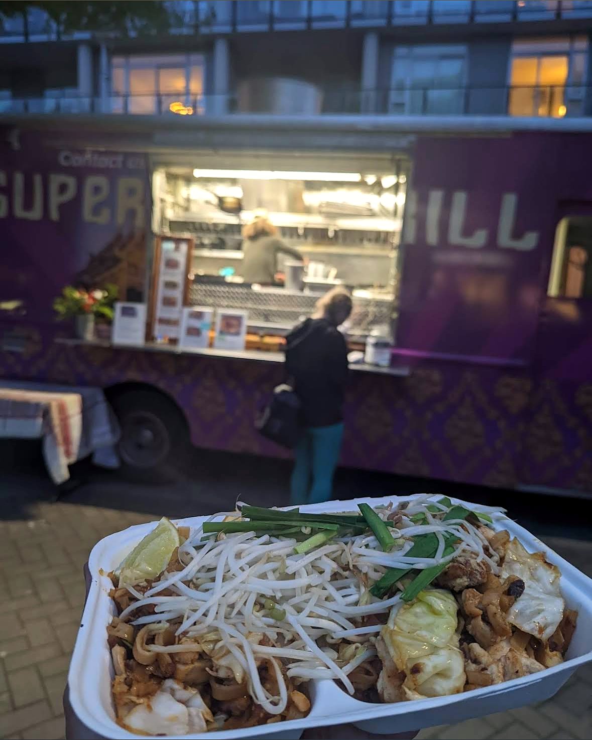 A tray of Pad Thai held up in front of the lit Super Thai truck at dusk